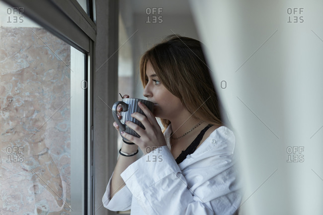 Young woman wearing oversized white blouse sipping coffee as she looks out window
