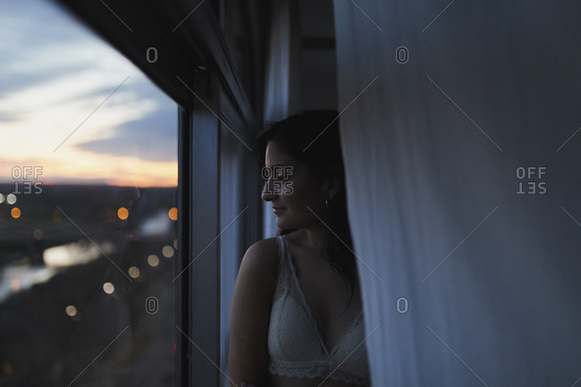 Woman in bra looking out high rise apartment window at dusk stock photo -  OFFSET