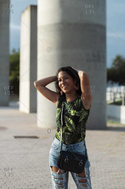 Beautiful young woman in stylish outfit touching hair and smiling while standing near gray wall on city street