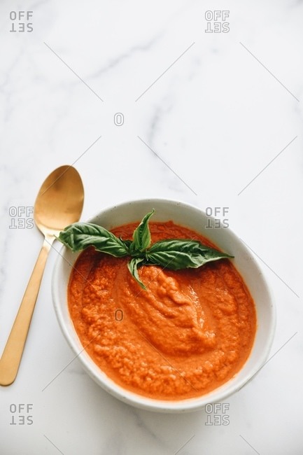 Top view of tomato soup with basil on white marble surface with gold utensils