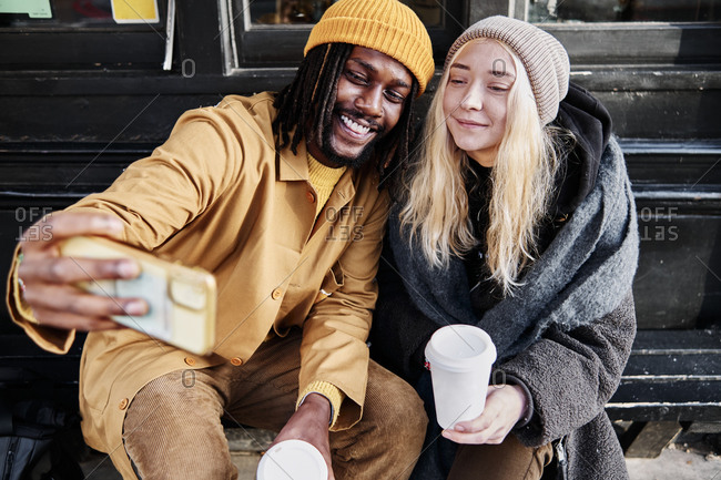 Cool biracial friends taking a selfie while sitting outside holding their cup of coffee