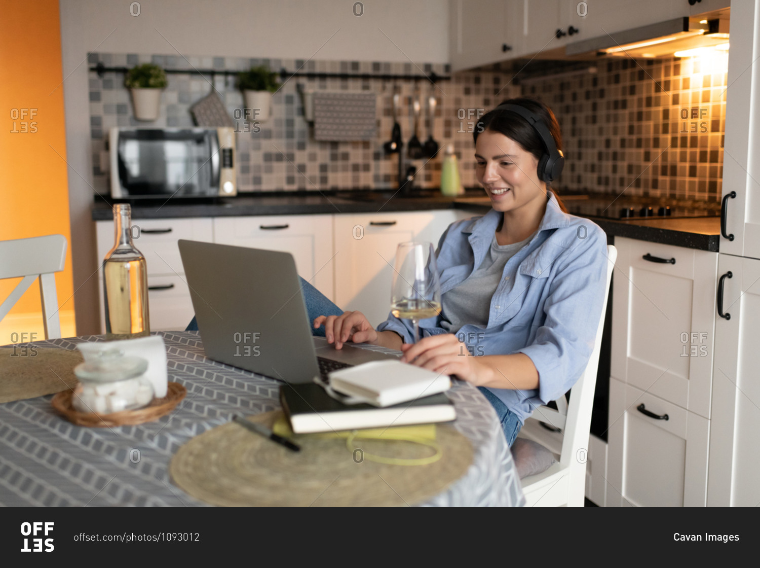 Happy woman sitting in kitchen with glass of wine and watching video on laptop