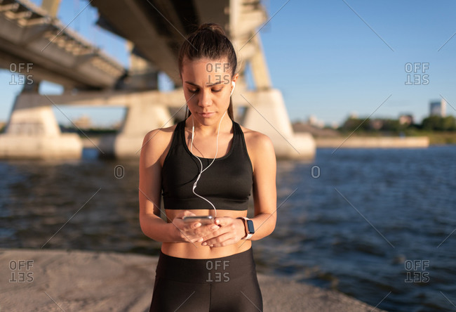 Slim sportswoman in headphones using phone on coast