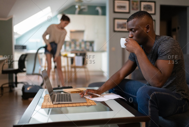 Black freelancer using laptop while girlfriend tidying living room