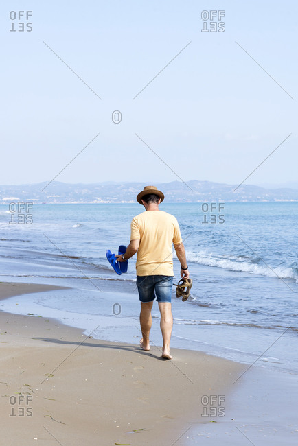 Rear view of man with hat, holding shoes walking barefoot on seashore