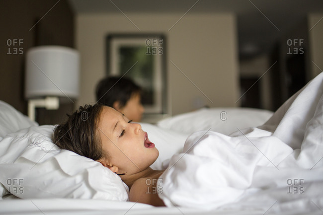 A little girl lays in a hotel room bed singing brother in background