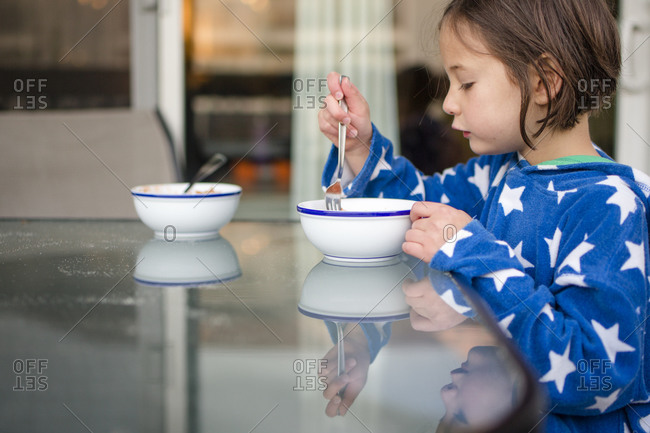 A small girl reflected in glass table eats breakfast outside