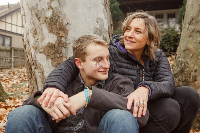 A smiling man sits against tree and wraps arm around teenage son