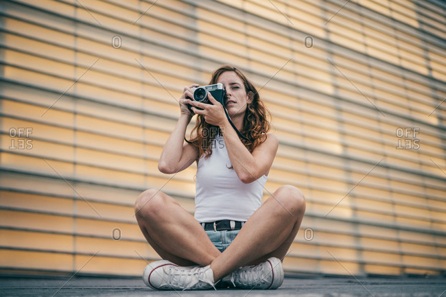 Young woman photographing through camera while sitting on footpath