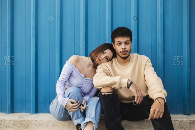 Girlfriend leaning on boyfriend's shoulder while sitting against blue wall