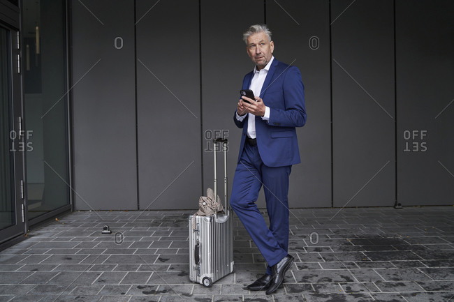 Businessman using mobile phone while standing with luggage on footpath