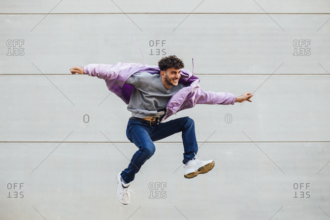 Carefree young man with arms outstretched jumping against gray wall