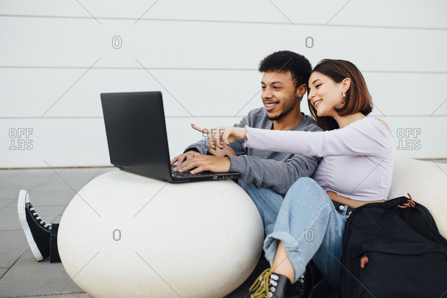 Smiling couple sitting and using laptop on white concrete ball