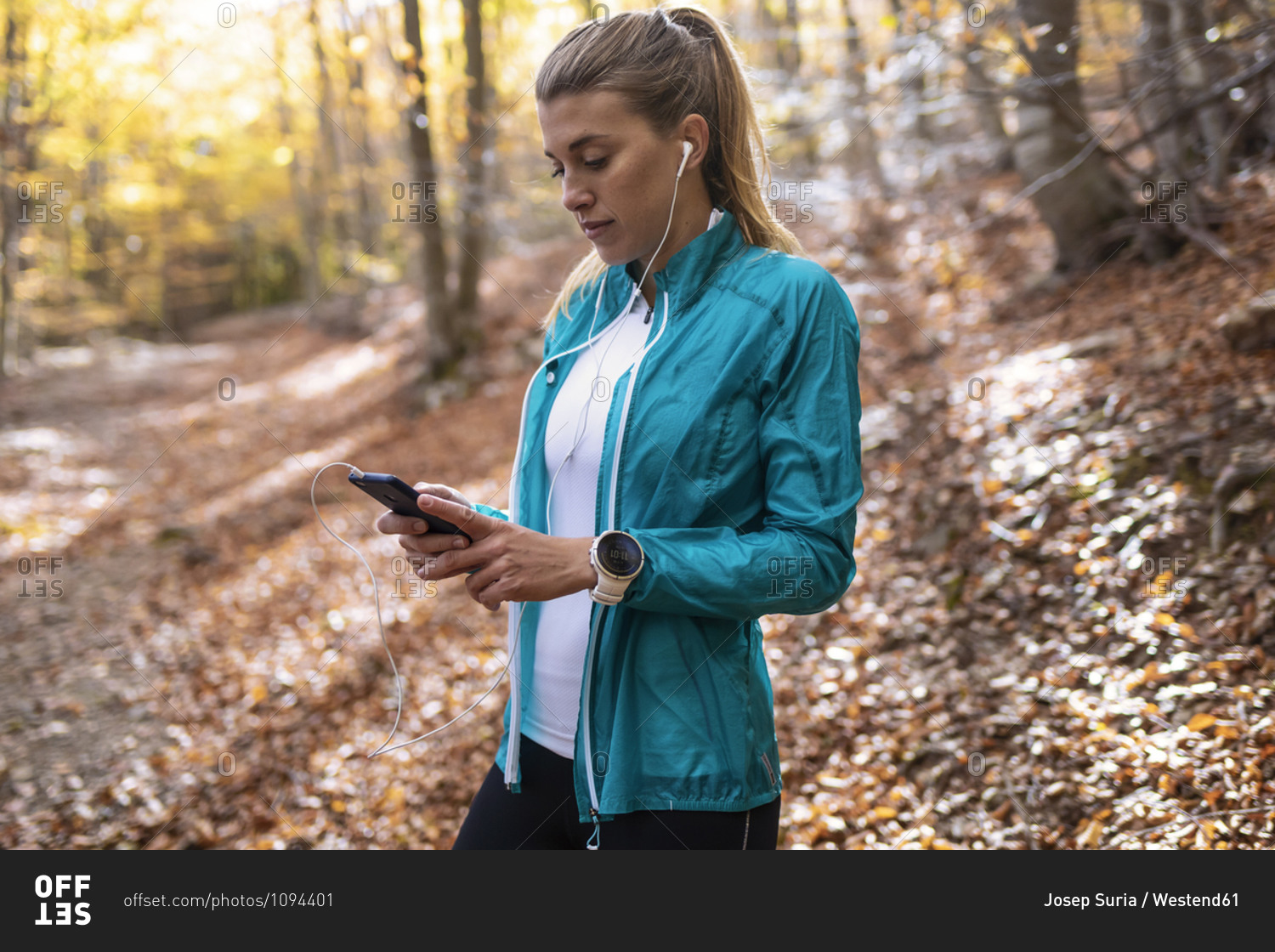 Sportswoman with in-ear headphones using mobile phone while standing in forest