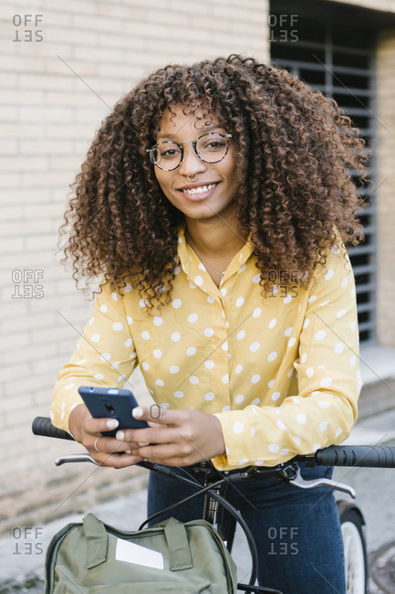 Smiling woman using mobile phone while standing with bicycle against wall