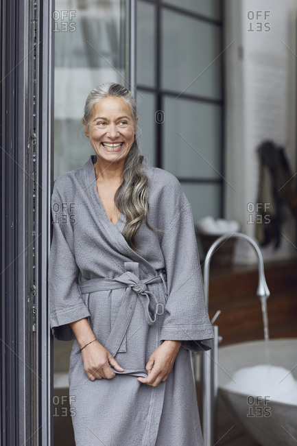 Smiling mature woman wearing bathrobe leaning by glass door in bathroom