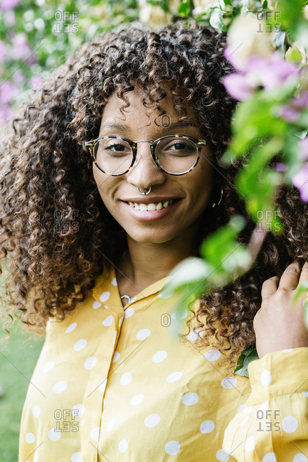 Curly hair woman smiling while standing by green wall at park