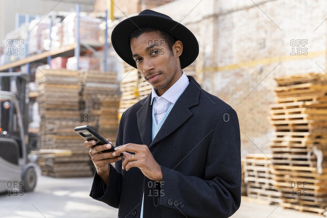 Fashionable man with cool attitude holding mobile phone in wood pallets warehouse
