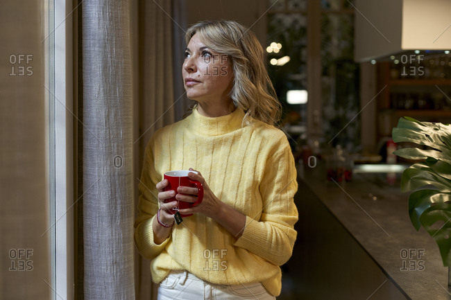 Fashionable mature woman looking through window while holding tea cup in kitchen