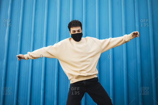 Carefree man wearing face mask jumping with arms outstretched against blue wall