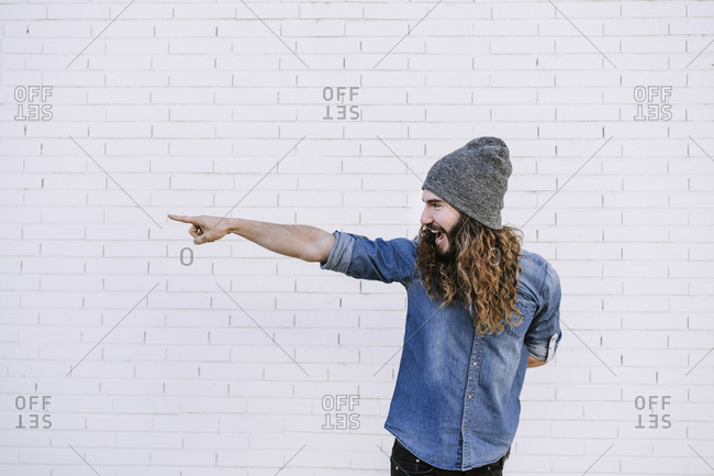 Happy young man in knit hat pointing while standing against brick wall