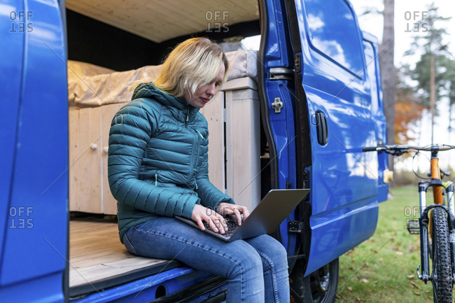 Blond woman working on laptop while sitting at door of camper van