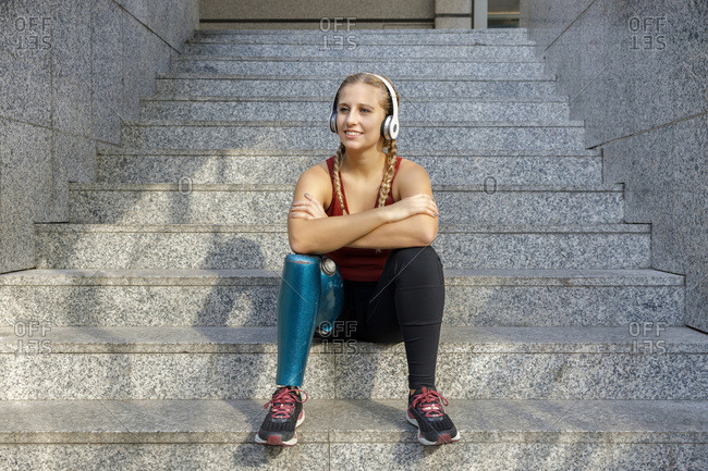 Front view of smiling female with bionic leg prosthesis sitting in street  and enjoying music in headphones on sunny day stock photo - OFFSET