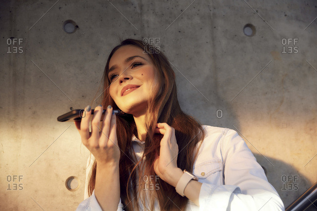 Smiling businesswoman hand in hair talking on mobile phone while standing against wall