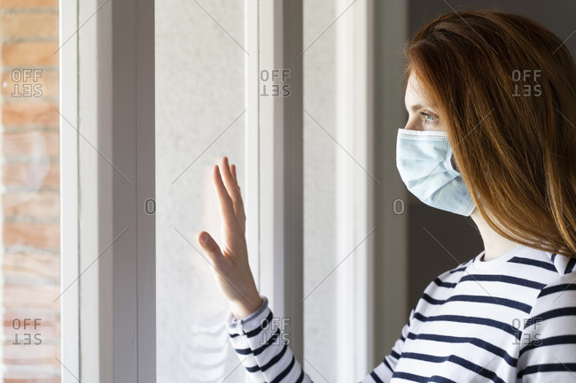 Redhead woman wearing protective face mask standing by window at home during COVID-19