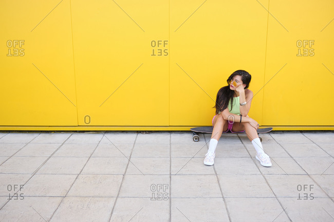 Portrait of beautiful girl sitting alone on longboard in front of yellow wall