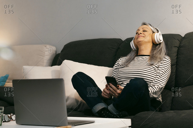 Smiling senior woman with mobile phone and laptop relaxing on sofa at home