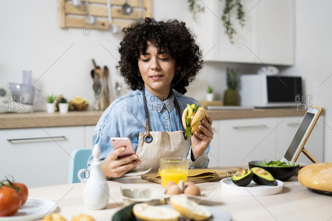 Portrait of young woman using smart phone at kitchen table with sandwich in hand