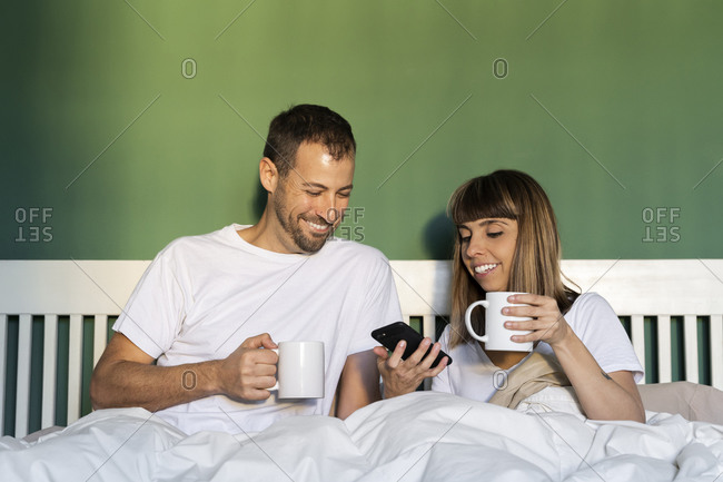 Smiling couple with coffee cup using mobile phone while sitting in bed at home