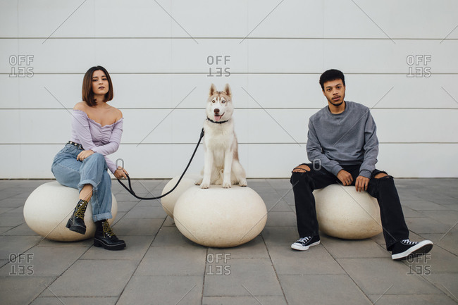 Boyfriend and girlfriend sitting by dog with social distance on white concrete ball against wall