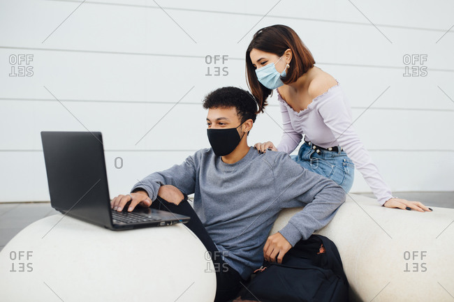Young couple wearing face mask using laptop while sitting by white concrete ball against wall