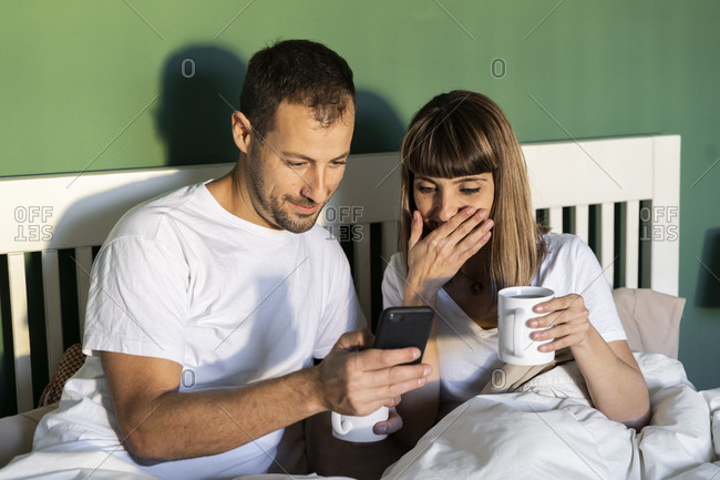 Boyfriend and girlfriend with coffee cup using mobile phone while sitting in bed at home