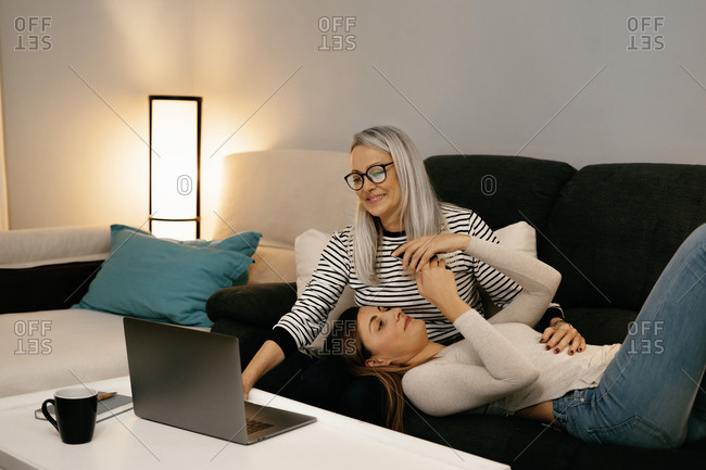 Daughter using mobile phone while lying on mother's lap sitting and using laptop at home