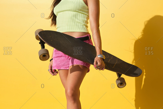 Mid section of young woman standing in front of yellow wall with longboard in hand