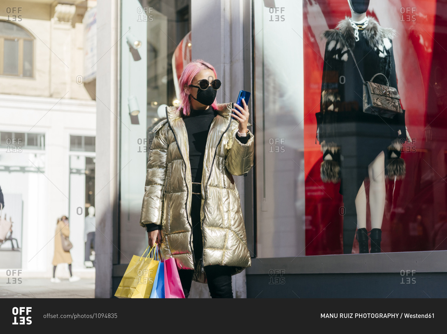 Fashionable woman with face mask carrying shopping bags while using mobile phone standing by store window