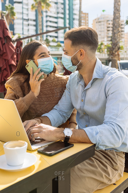Unrecognizable young ethnic female entrepreneur in stylish clothes and medical mask talking on smartphone and looking at boyfriend working on laptop in street cafe during coronavirus