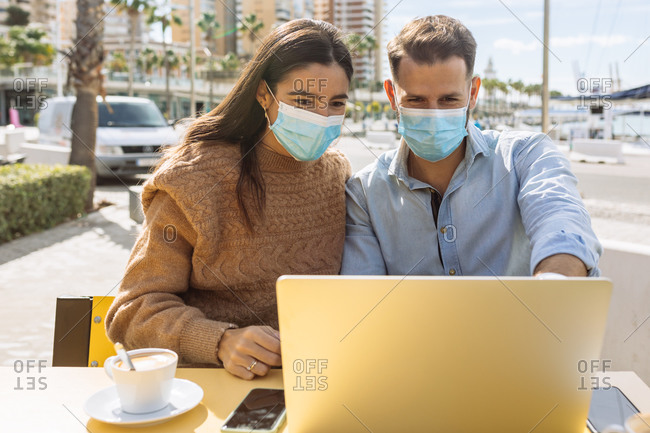 Unrecognizable young ethnic female entrepreneur in stylish clothes and medical mask talking on smartphone and looking at boyfriend working on laptop in street cafe during coronavirus