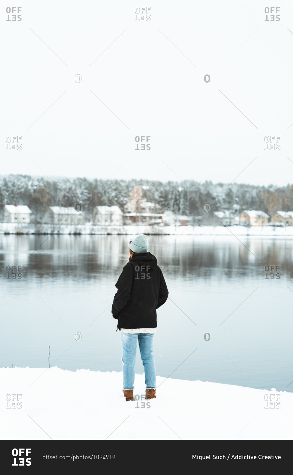 Back view of female traveler standing on shore of pond and admiring scenery of small village in winter forest