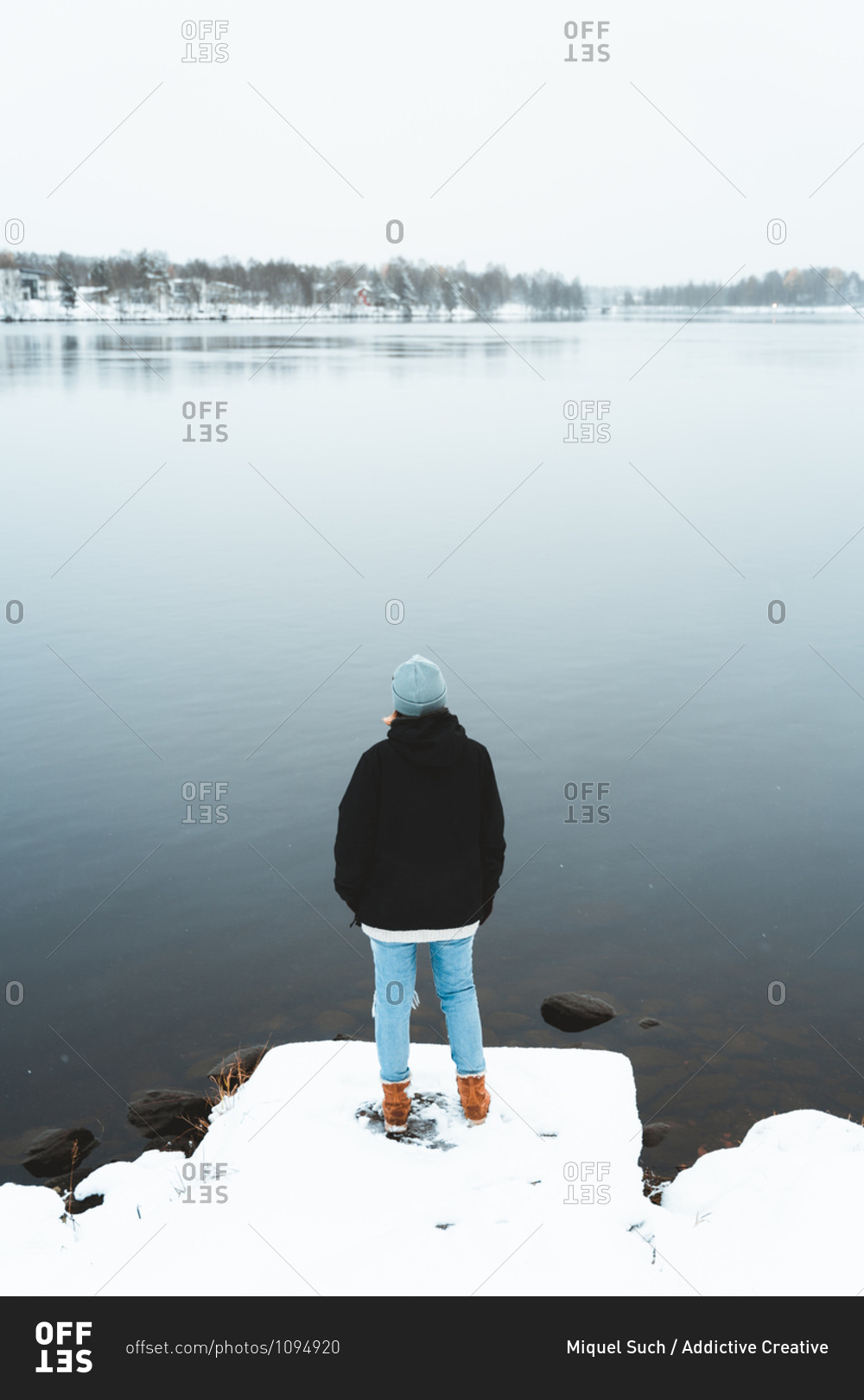 Back view of female traveler standing on shore of pond and admiring scenery of small village in winter forest