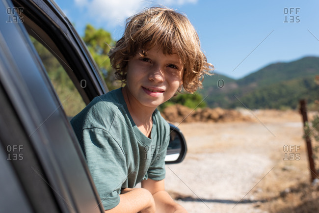 Side view of preteen boy with curly hair sitting in automobile looking out a window of while enjoying summer adventure in mountainous land