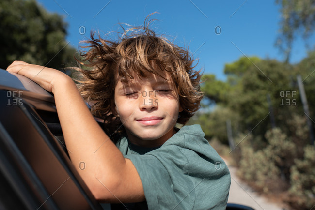 Side view of preteen boy with curly hair sitting in open window of automobile with eyes close while enjoying summer adventure in mountainous land