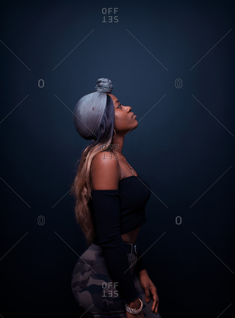 Side view of attractive young African female model in stylish outfit with bare shoulders and traditional headwear standing against dark background looking away