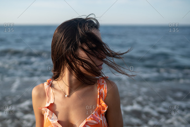 Side view of dreamy young tanned ethnic female traveler in stylish swimwear and sunglasses recreating on rocky seashore on windy day and looking away