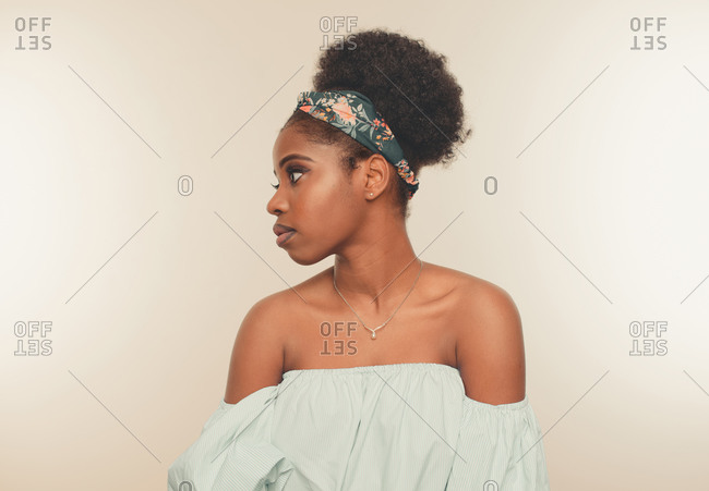 Young slim African American female model with Afro hair bun and headband wearing stylish blue crop top and jeans standing against beige background looking away
