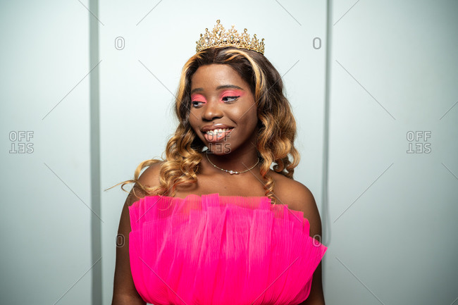 Self assured young black lady with long curly hair and makeup in pink fashionable dress and golden crown looking at camera during beauty contest