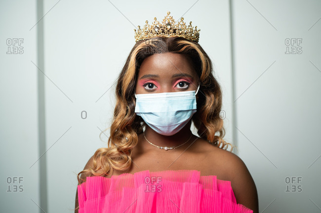 Unrecognizable young African American female with long wavy hair in crown on head and medical mask standing against white wall and looking at camera during coronavirus